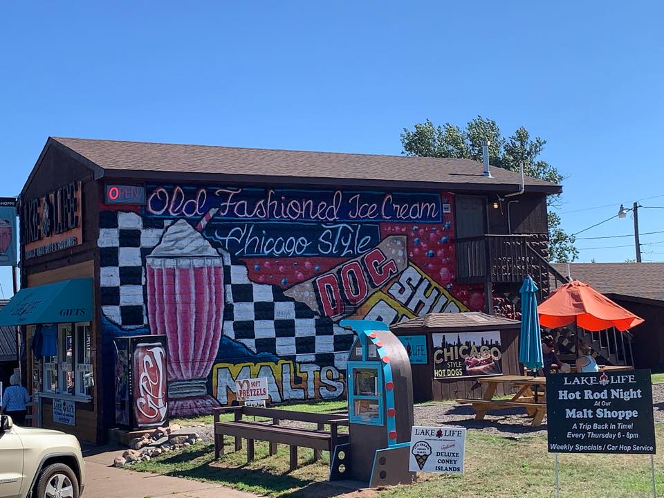 A mural on a wooden building features a large pink milkshake, checkerboard patterns, and the words "Old Fashioned Ice Cream Chicago Style"—a fun spot for summer activities in Superior, Wisconsin with hot dogs, malts, gifts, and hot rod night.
