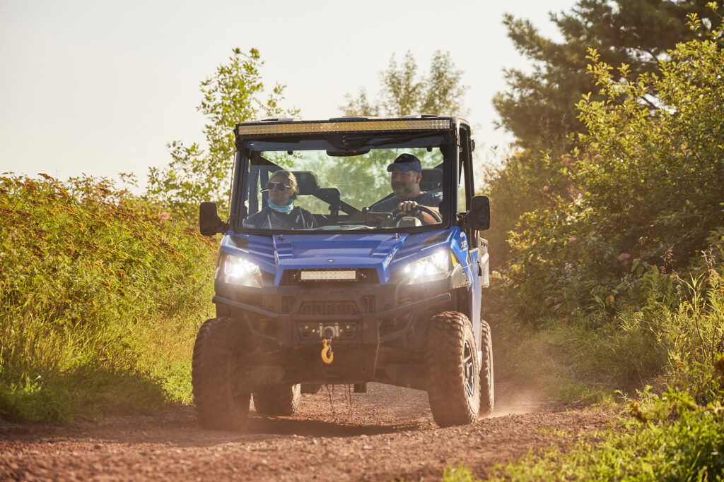 A woman stands in front of a large window, smiling, with a city skyline and tall buildings behind her under a blue sky with scattered clouds—perfect weather for summer activities in Superior Wisconsin. She is wearing a dark suit and white blouse.