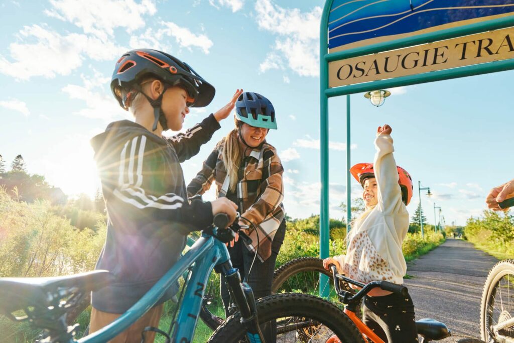 Three children wearing helmets and casual clothes smile and high-five while holding their bikes on a sunlit trail under a blue sign reading “OSAUGIE TRAI.” Trees and a clear sky are visible in the background.
