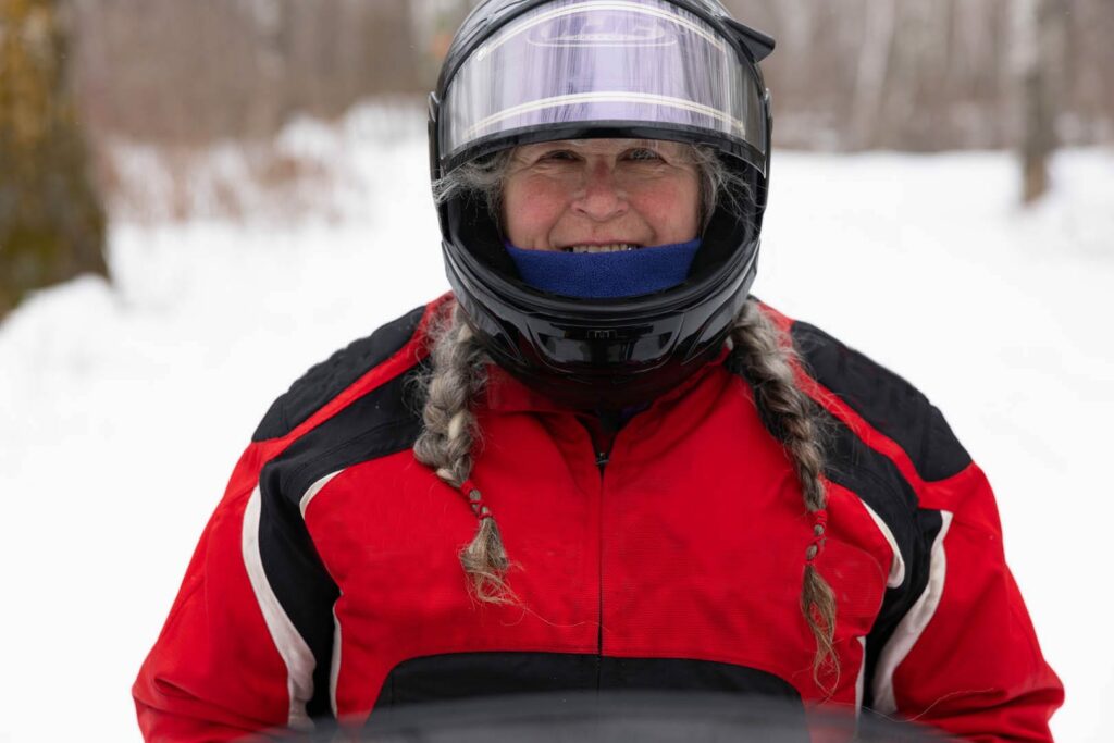 A person with gray braided hair, wearing a red and black jacket and a black helmet with a clear visor, stands outdoors in a snowy, wooded area, smiling at the camera.