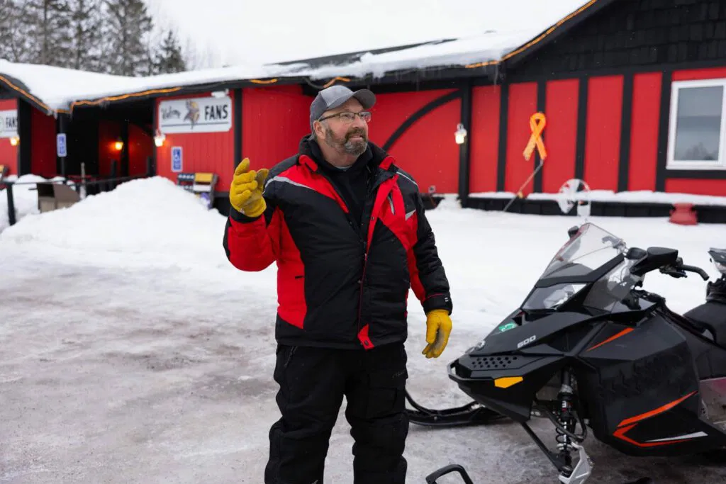 A man in a red and black winter jacket and yellow gloves stands outside in the snow next to a snowmobile, with a red and black building in the background.