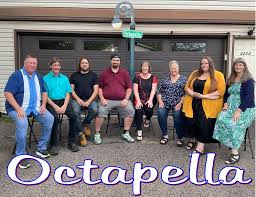 Eight people sit in a row outdoors in front of a garage, posing for a group photo under a street lamp. The word "Octapella" is displayed at the bottom of the image in large script font.