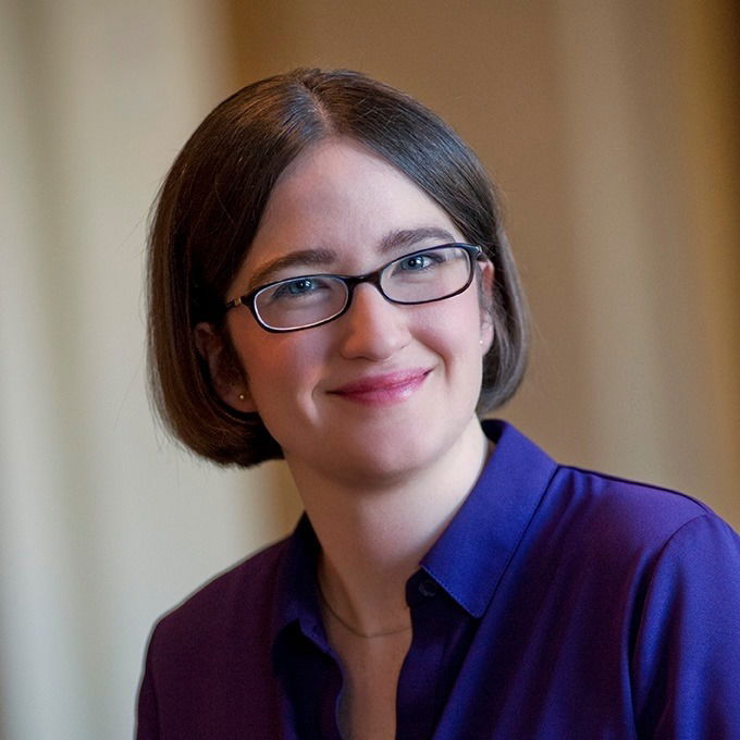 A woman with straight brown hair, glasses, and a purple blouse smiles softly at the camera against a softly blurred neutral background.