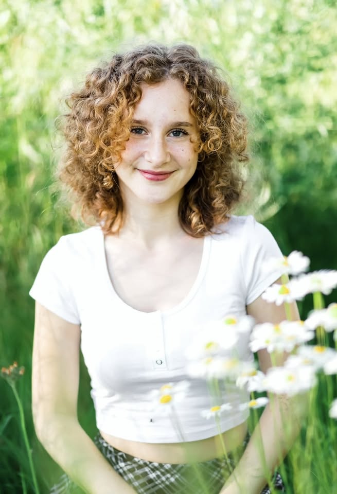 A young woman with curly red hair, wearing a white short-sleeved top, smiles while sitting outdoors among green grass and white daisies on a sunny day.