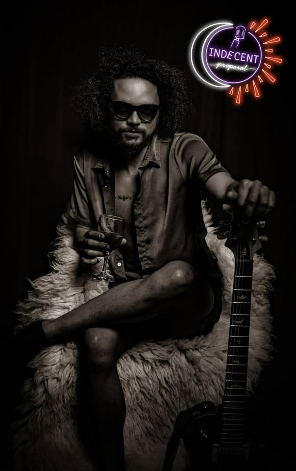 A man with curly hair and sunglasses sits on a furry chair, holding a drink and resting his hand on an electric guitar. A neon sign reading "Indecent Proposal" glows in the background.