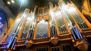 A close-up view of a large pipe organ with numerous shiny metal pipes and ornate wooden casing, illuminated by bright indoor lighting in what appears to be a church or cathedral.