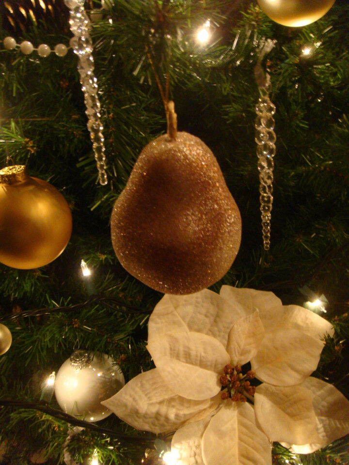 A glittery pear-shaped ornament hangs on a Christmas tree decorated with gold and white baubles, crystal garlands, and a large white poinsettia flower. Warm lights illuminate the festive scene.