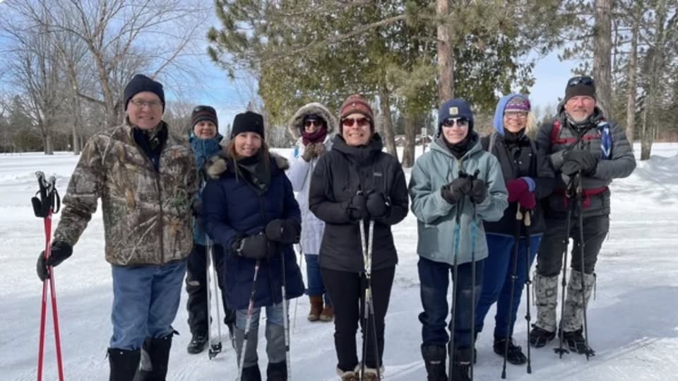 A group of eight people dressed in winter clothing stand outdoors on a snowy trail with ski poles, surrounded by trees on a sunny day.