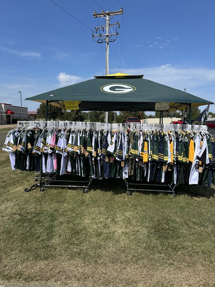 A large rack of Green Bay Packers jerseys is displayed on a grassy lawn under a tent with a Packers logo, on a sunny day. Power lines and a few buildings are visible in the background.