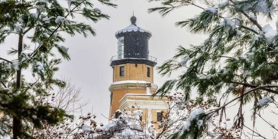 A tan brick lighthouse with a black lantern stands surrounded by snow-covered pine branches and trees, under a cloudy winter sky.