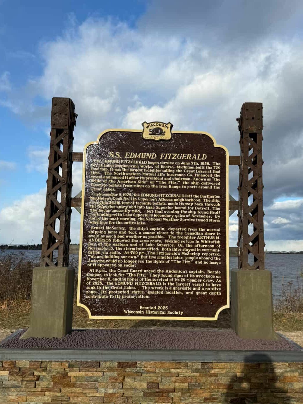 A historical marker for the S.S. Edmund Fitzgerald stands outdoors near a body of water, with cloudy skies above and dry grass in the background. The sign details the ship’s history.