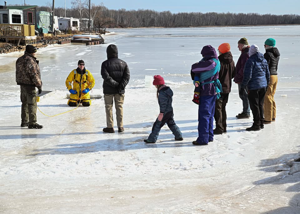 A group of people stands on an icy lakeshore, watching a person in a yellow rescue suit kneeling on the ice, holding a rope. Trees, trailers, and a partially frozen lake are visible in the background.