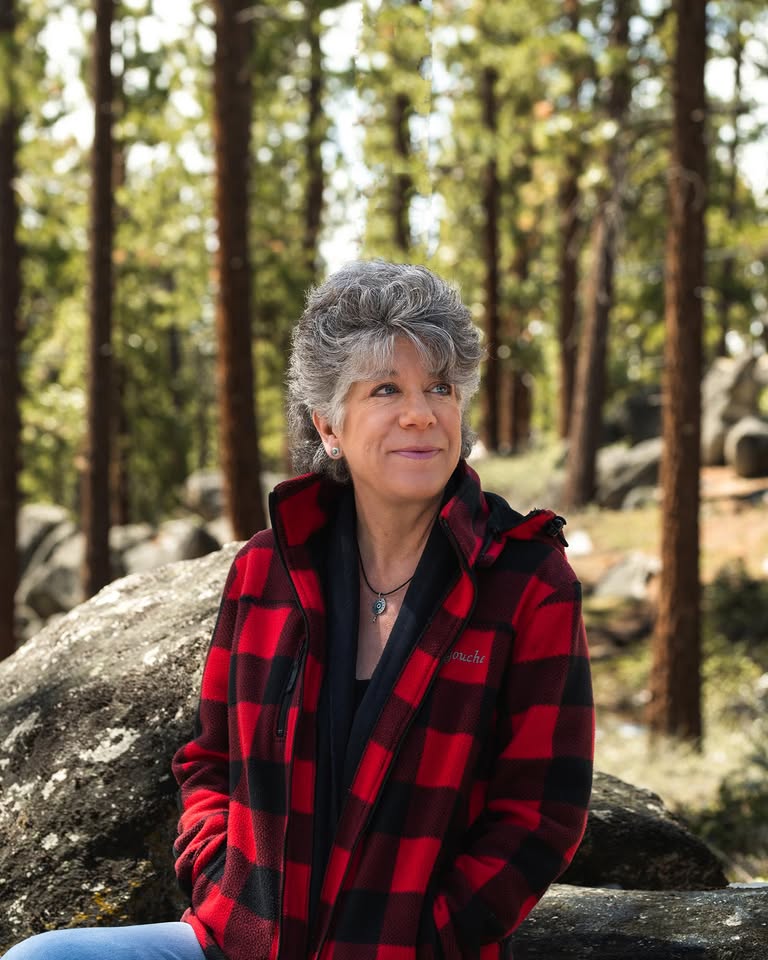 A woman with short gray hair wearing a red and black plaid jacket sits on a large rock in a sunlit forest, smiling thoughtfully with pine trees and boulders in the background.