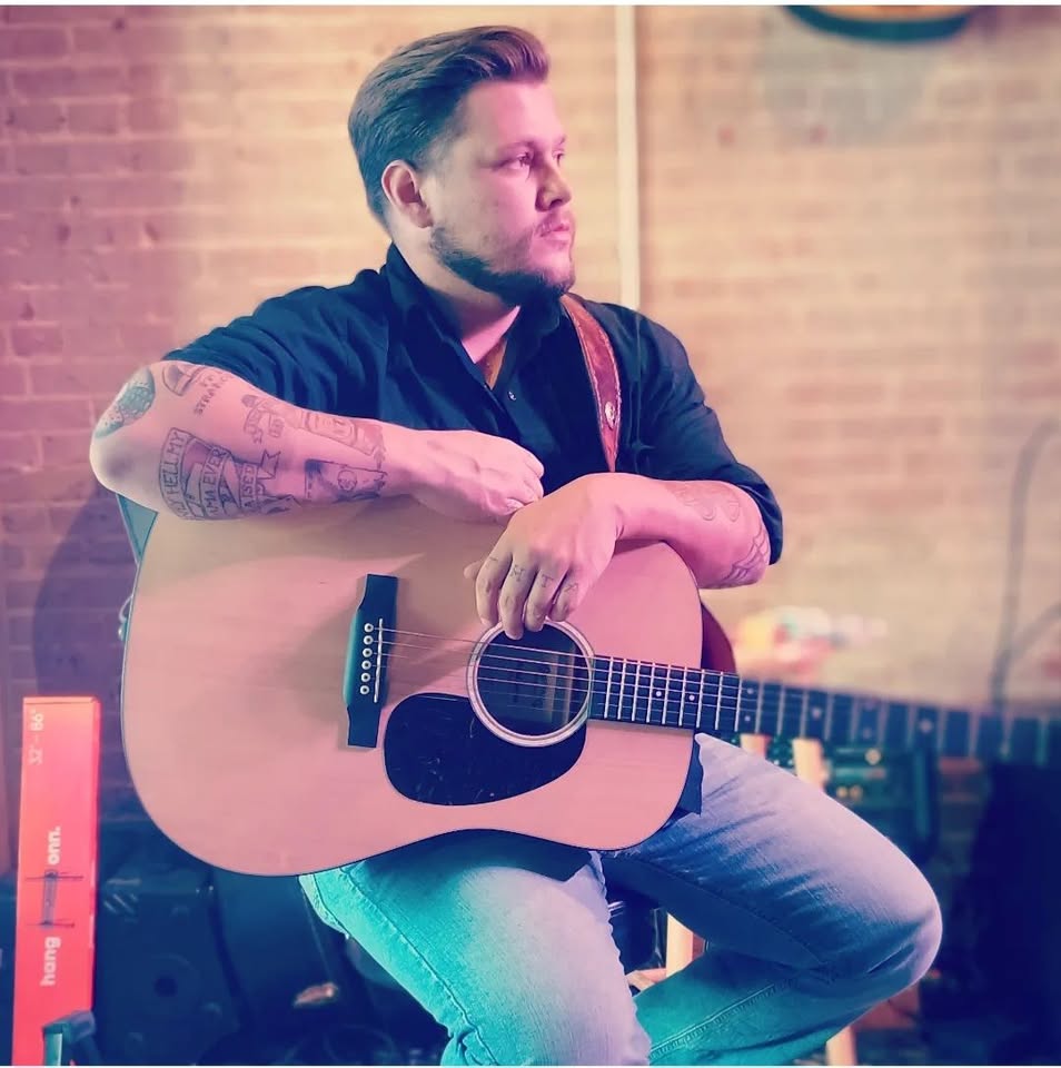 A man with tattoos sits holding an acoustic guitar, wearing a dark shirt and jeans. He looks to the side in a softly lit room with exposed brick walls and a relaxed atmosphere.