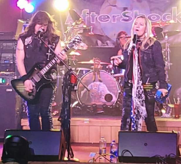 Two musicians perform on stage; the man on the left plays an electric guitar and the woman on the right sings into a microphone. Drums and stage lights are visible in the background with a banner reading "AfterShock.