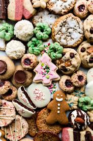 An assortment of festive holiday cookies, including frosted sugar cookies, gingerbread, peanut butter cups, thumbprint cookies, and cookies with colorful sprinkles and icing, arranged closely together.