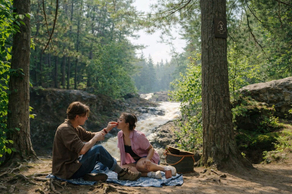 A man and woman sit on a blanket in a forest near a waterfall, sharing food—a perfect scene for weekend getaways in Wisconsin. The woman is being fed by the man, with a picnic basket nearby as sunlight filters through the trees.