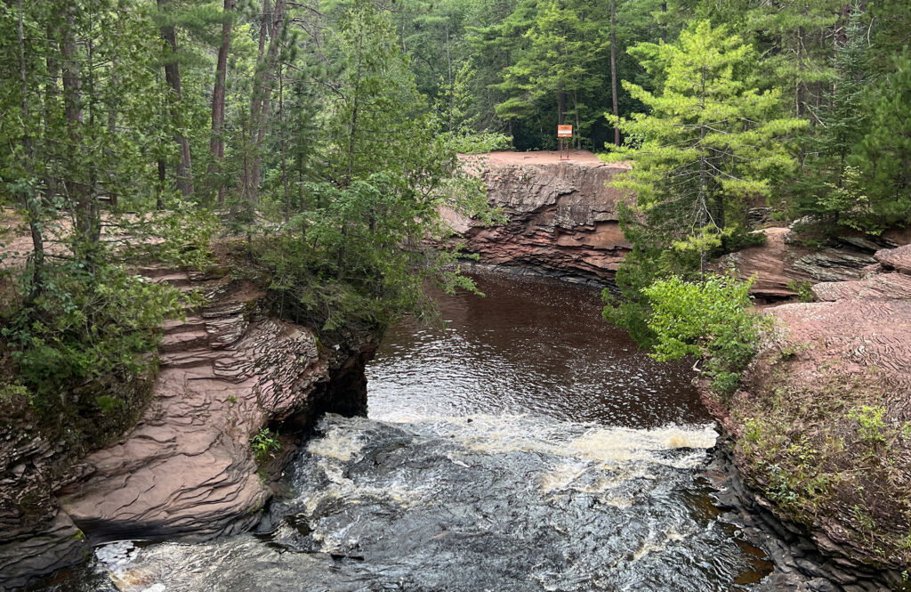 A river flows through a rocky, forested landscape with reddish stone cliffs and dense green trees—typical scenery of parks in Superior, Wisconsin. Water cascades over rocks in the foreground, with a warning sign visible on the cliff above.