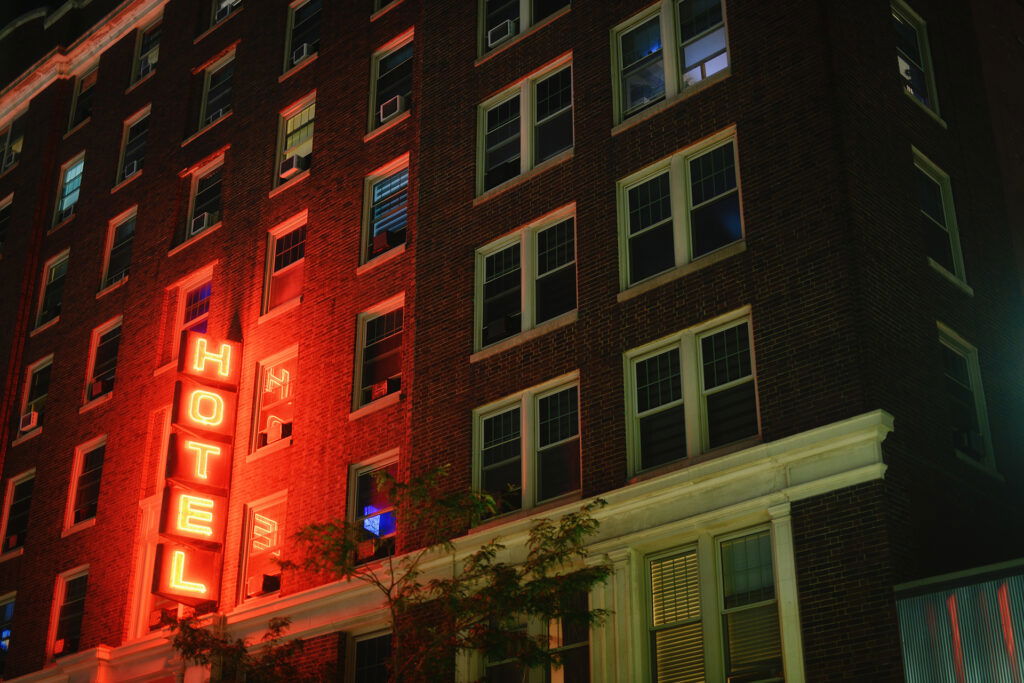 A neon red "HOTEL" sign glows outside a multi-story brick building at night, perfect for a memorable Wisconsin Family Vacation. Warm light spills from the windows, and a tree stands below, adding to the inviting atmosphere.