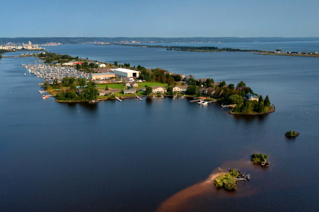 Aerial view of a small peninsula with buildings, trees, and a marina jutting into a large body of water—perfect for water activities in Superior, Wisconsin—with a distant city skyline visible across the bay under a clear blue sky.