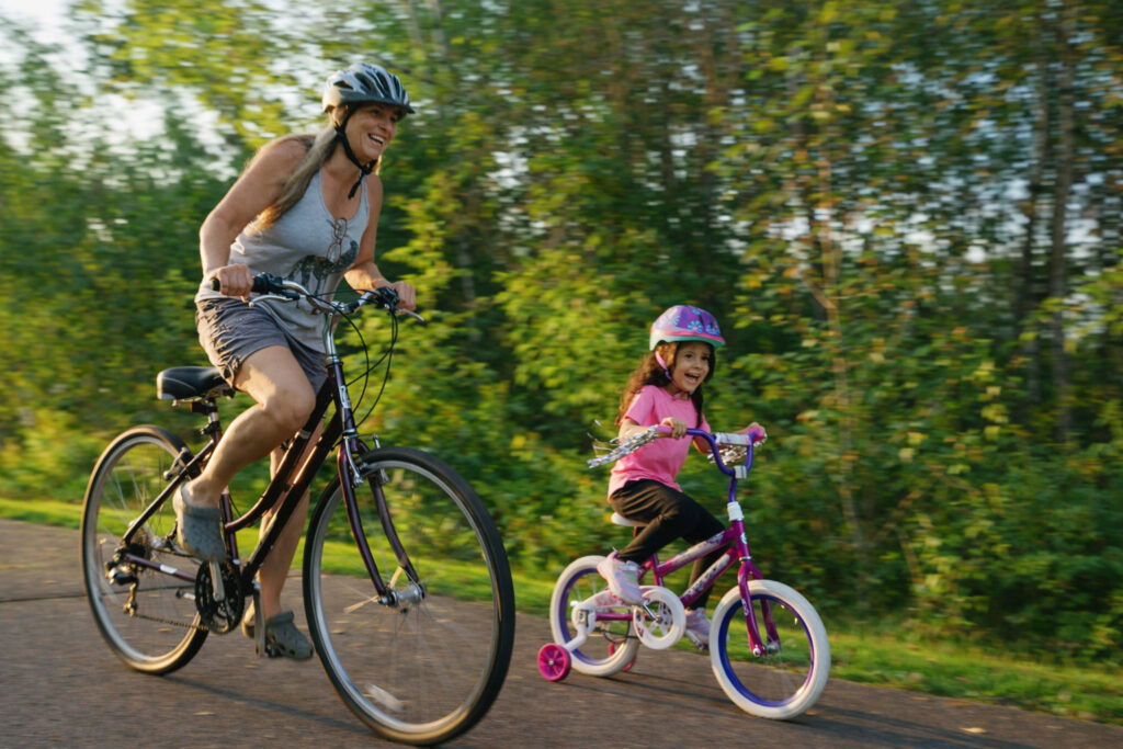 A woman and a young girl, both in helmets, ride bicycles together on a sunny park path surrounded by green trees—perfect for those seeking weekend getaways in Wisconsin where families can enjoy the outdoors and share smiles.
