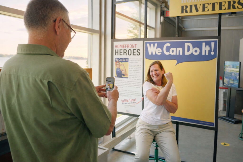 A woman poses in front of a “We Can Do It!” sign, flexing her arm like Rosie the Riveter, while a man snaps her photo—capturing an inspiring moment during one of their weekend getaways in Wisconsin’s museums.