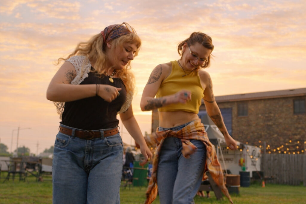 Two women with tattoos joyfully dance together outdoors at sunset in Superior, Wisconsin. One wears a black top and glasses, the other a yellow crop top; both are smiling amid string lights and a brick building—perfect for your list of things to do in Superior Wisconsin.