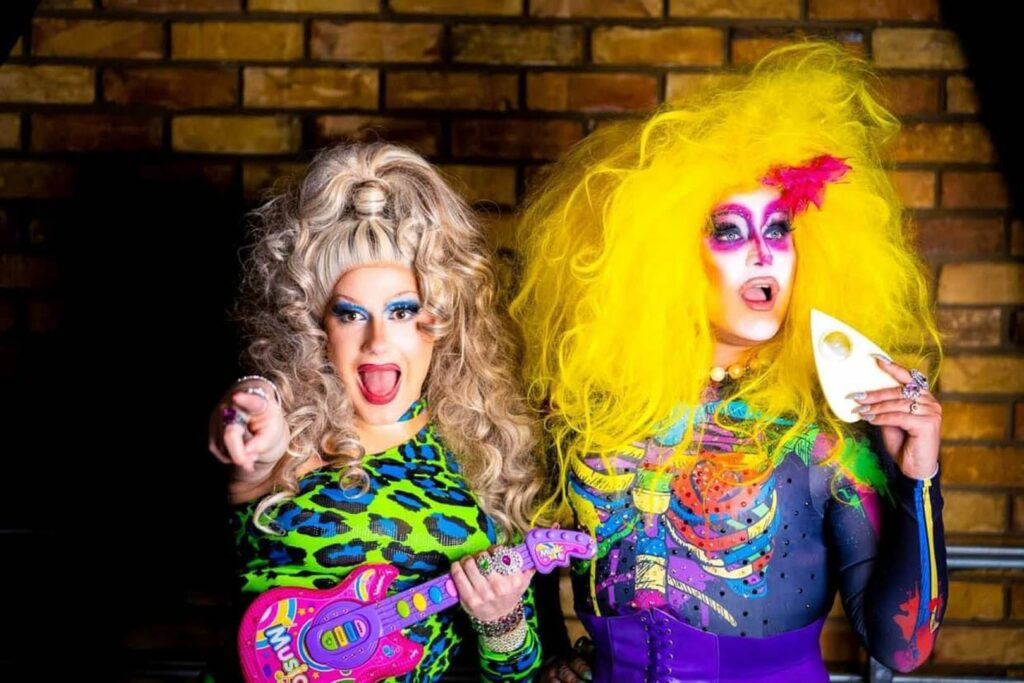 Two drag queens stand in front of a brick wall, channeling the energy of weekend getaways in Wisconsin. One wears a colorful animal print outfit, holds a toy guitar, and points forward; the other rocks a neon yellow wig, bold face paint, and waves a white paper fan.