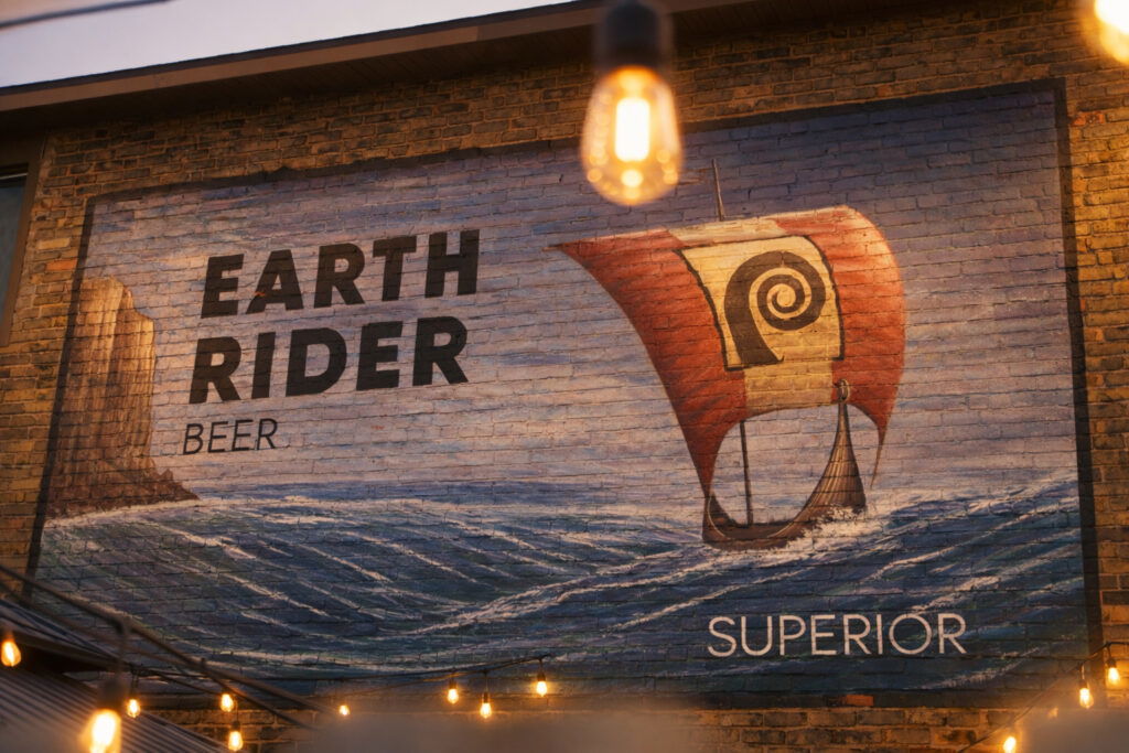 A mural on a brick wall shows a Viking ship with a red sail on the ocean, next to text reading "EARTH RIDER BEER SUPERIOR." String lights and a hanging bulb set the scene for fall activities in Superior, Wisconsin.