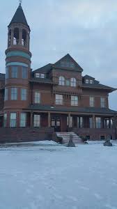 A large, historic-looking brick mansion with a tall, round turret stands on snow-covered ground under a cloudy sky.