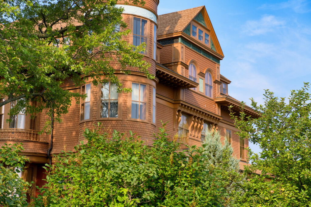 A large, historic brick house with ornate architectural details and multiple windows, partly obscured by green trees and shrubs, under a clear blue sky—reminiscent of the charming Museums in Superior Wisconsin.