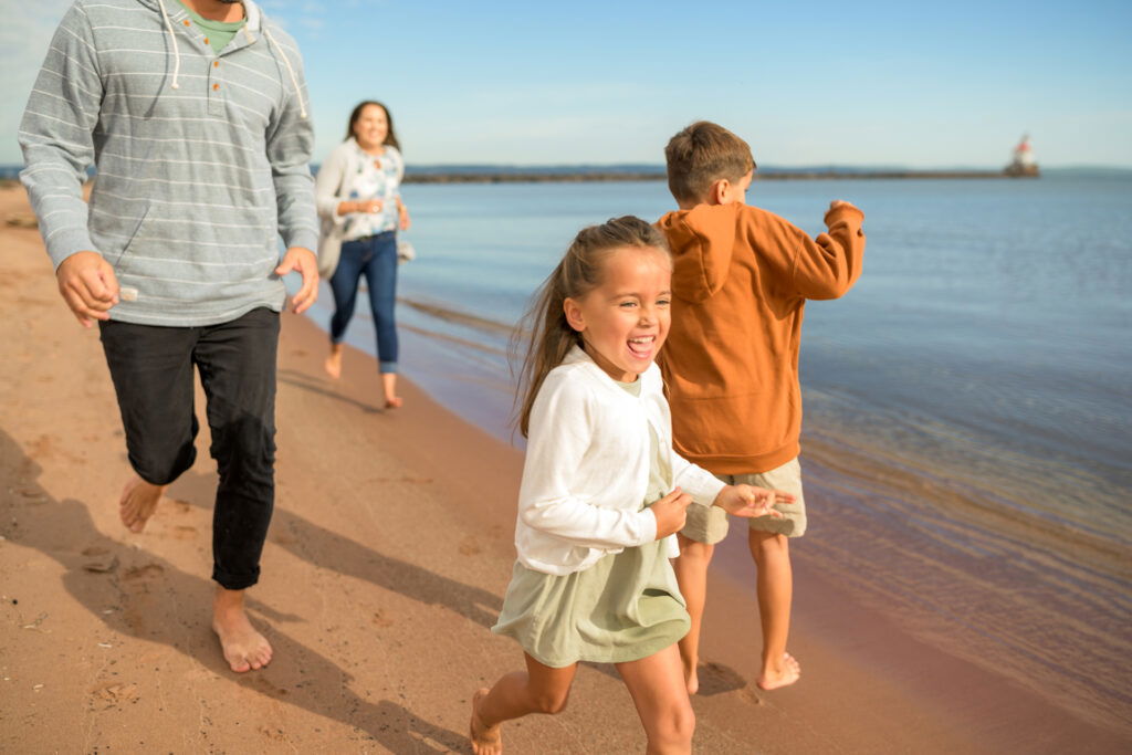 Two children and two adults run and laugh along the shoreline of a sandy beach on a sunny day—classic fun that makes Wisconsin family vacations unforgettable, with calm water and a clear blue sky in the background.