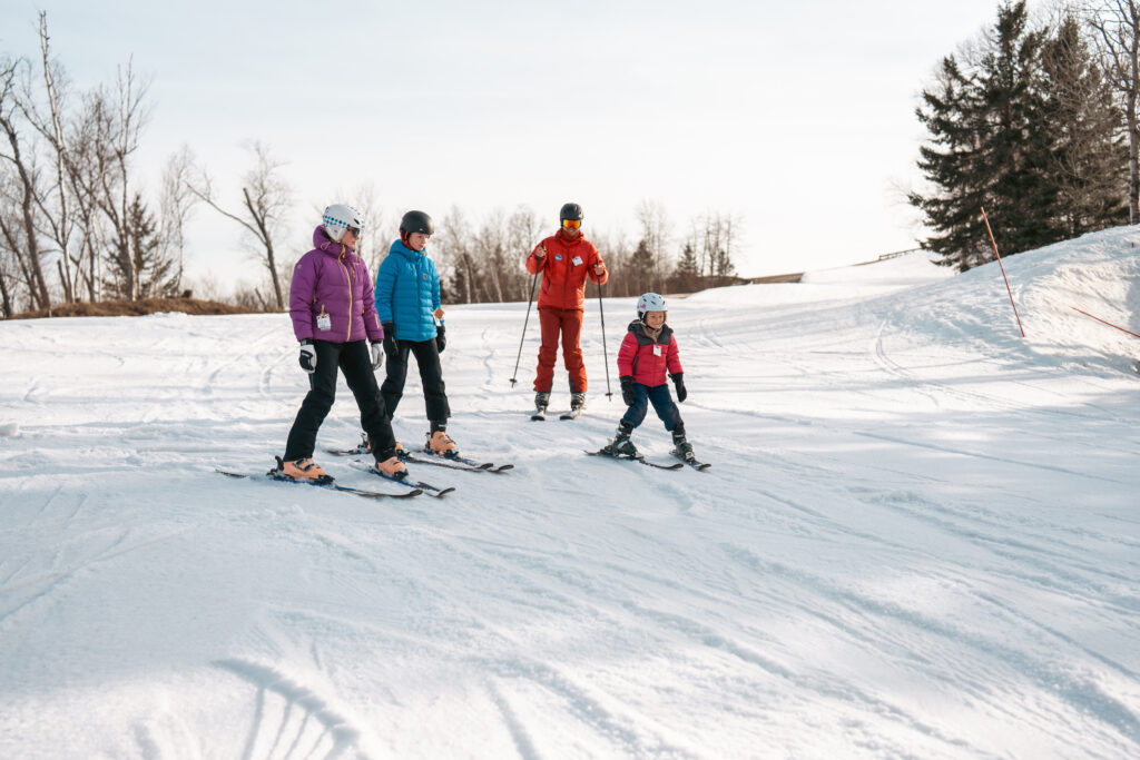 Four people, including a child, ski on a snowy slope—a fun winter activity and one of the exciting things to do in Superior Wisconsin. An instructor in red guides them as they head downhill, with helmets and jackets against a backdrop of leafless trees and evergreens.