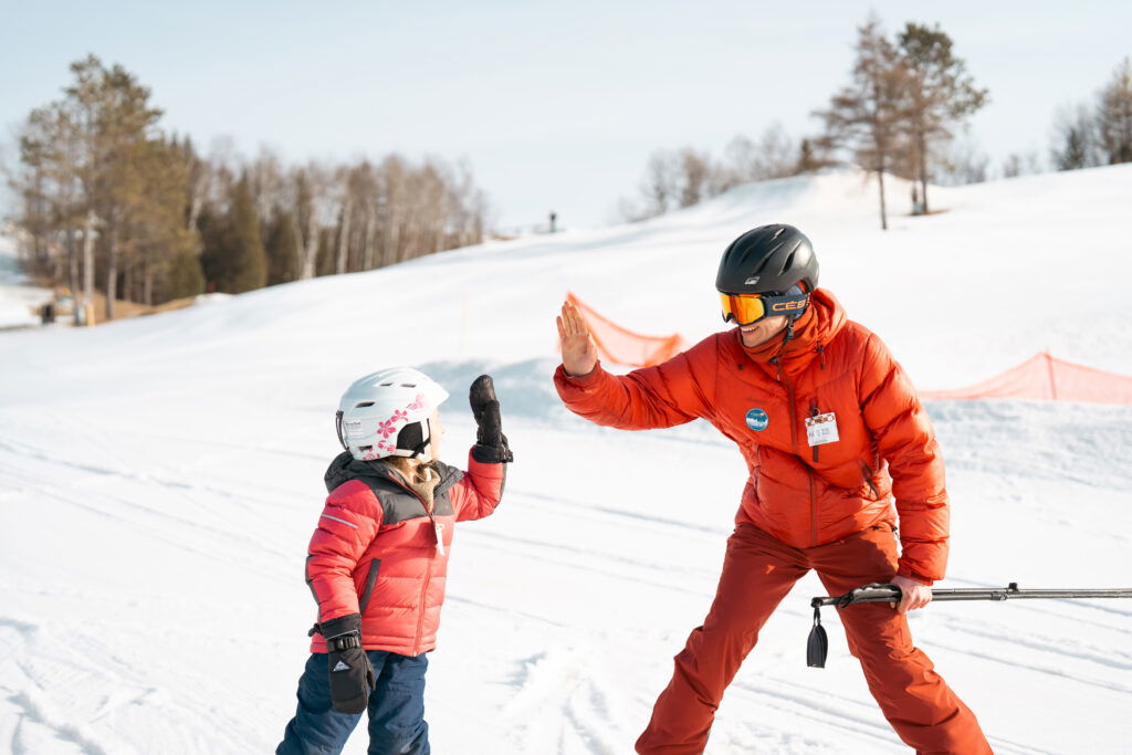 An adult and a child wearing helmets and red jackets give each other a high-five on a snowy ski slope in Wisconsin, with trees and orange safety barriers in the background—perfect for weekend getaways in Wisconsin.