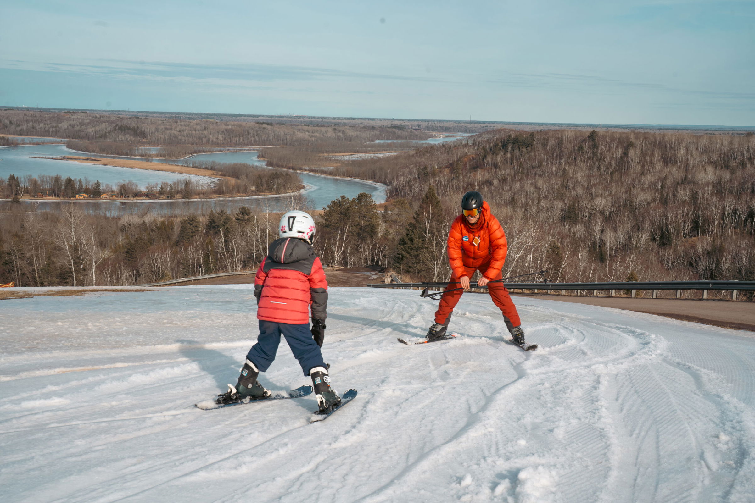 An adult and a child, both in helmets and red jackets, enjoy skiing in Superior Wisconsin as they glide down a snowy slope with scenic views of winding rivers and forested hills in the background.
