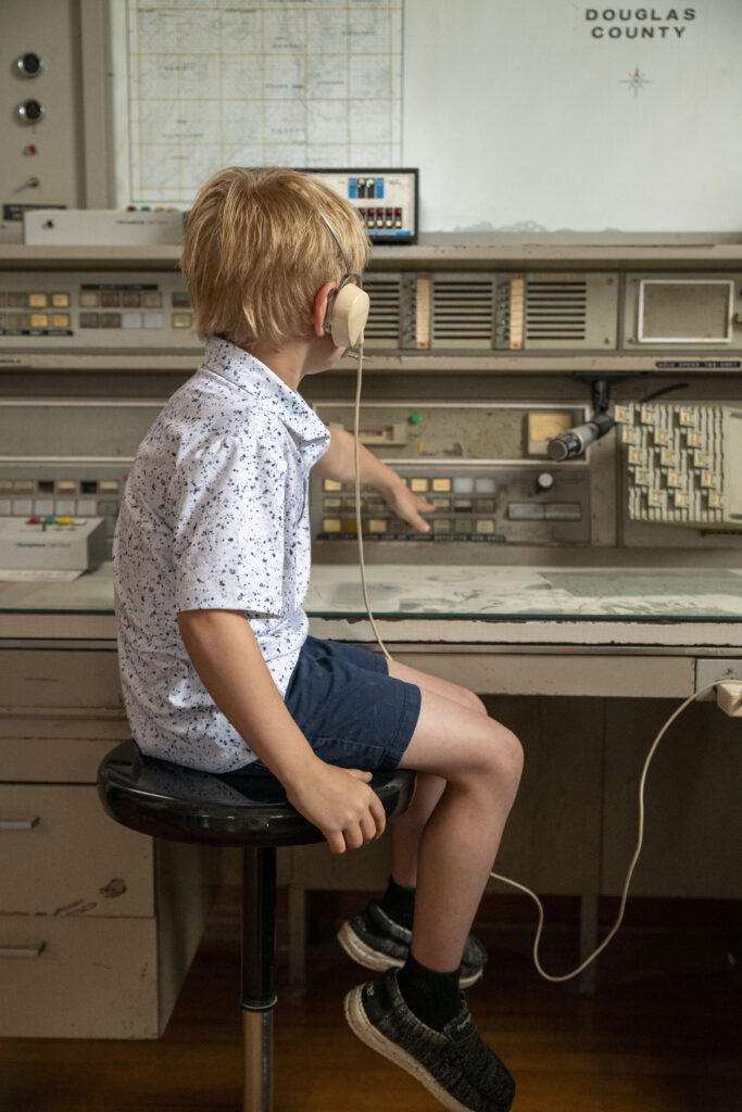A young boy wearing headphones sits on a stool and points at buttons on an old-fashioned control panel in front of a large map labeled "Douglas County"—a glimpse into interactive exhibits at museums in Superior, Wisconsin.