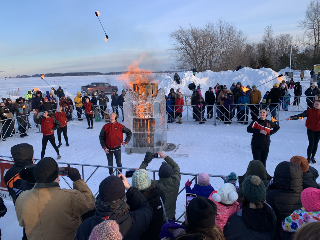 A crowd watches jugglers toss flaming clubs in front of a blazing ice sculpture at the snowy Lake Superior Ice Festival, with people bundled up in coats and hats enjoying the winter spectacle.