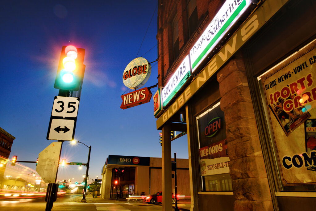 A city street corner at dusk with a green traffic light, speed limit sign, and a Globe News storefront with neon signs—evoking the lively atmosphere of a Wisconsin Family Vacation. Cars and buildings are visible under a deep blue sky.