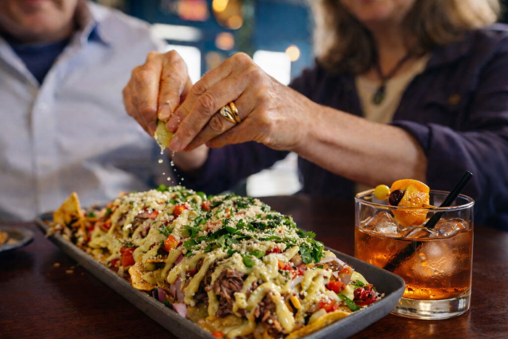 Two people sit at a table; one squeezes a lime over nachos topped with meat, cheese, and vegetables. A cocktail with an orange garnish sits nearby—perfect fuel for planning weekend getaways in Wisconsin.