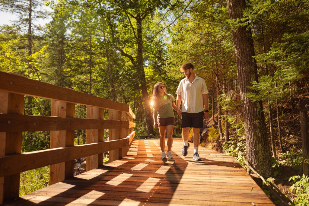A couple walks hand in hand on a sunlit wooden bridge surrounded by lush green trees in a forest, enjoying one of the free activities and attractions in Superior, Wisconsin.