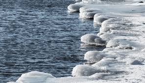 Chunks of ice and snow cover the shoreline, with partially frozen water and rounded icy formations extending into the lake or river. The surface shows a contrast between the frozen edge and open water.