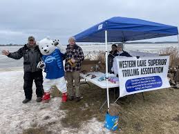Three people stand by a tent near a snowy lake; one is dressed in a polar bear mascot costume. A banner reads "Western Lake Superior Trolling Association." A table holds papers and supplies under the tent.