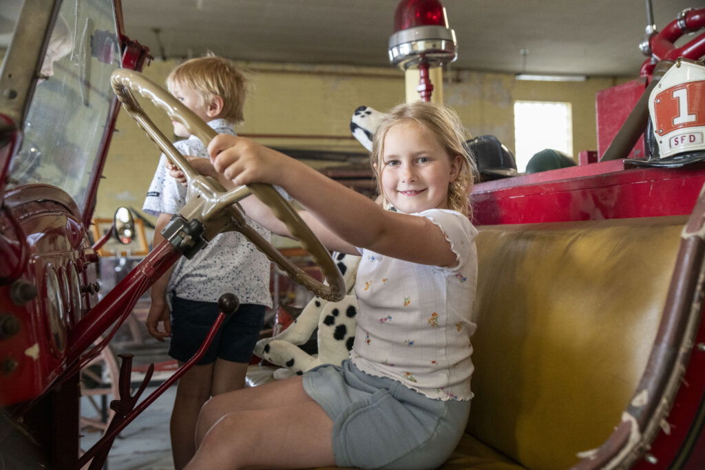 A young girl smiles while sitting in the driver's seat of a vintage red fire truck—an adventure perfect for weekend getaways in Wisconsin. A boy stands beside her, and a stuffed Dalmatian toy is in the background.