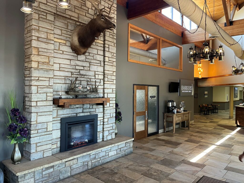 A lodge lobby with a stone fireplace, mounted deer head, floral arrangements, and a wooden mantel welcomes guests. A coffee station and "Barkers Island Inn" sign hint at Lake Superior Ice Festival fun beyond the tiled floors and large windows.