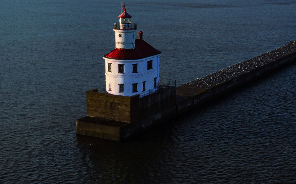 A white lighthouse with a red roof sits on a concrete pier extending into calm, dark blue water, with seagulls lining the pier—a picture-perfect scene for your Wisconsin family vacation.