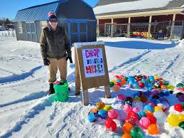 A person in winter clothing stands outside on snow near a sign that says “Drop Balloons Here!” Colorful balloons are scattered on the ground, with buildings and a fence in the background.