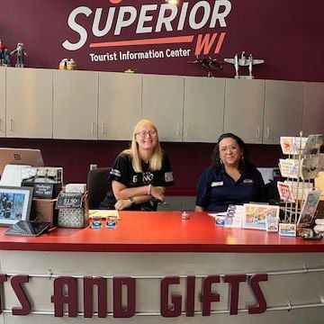 Two women smile behind the counter at the Superior, WI Tourist Information Center, ready to help you plan weekend getaways in Wisconsin. Brochures, a computer, and gift items decorate the desk with maroon walls in the background.