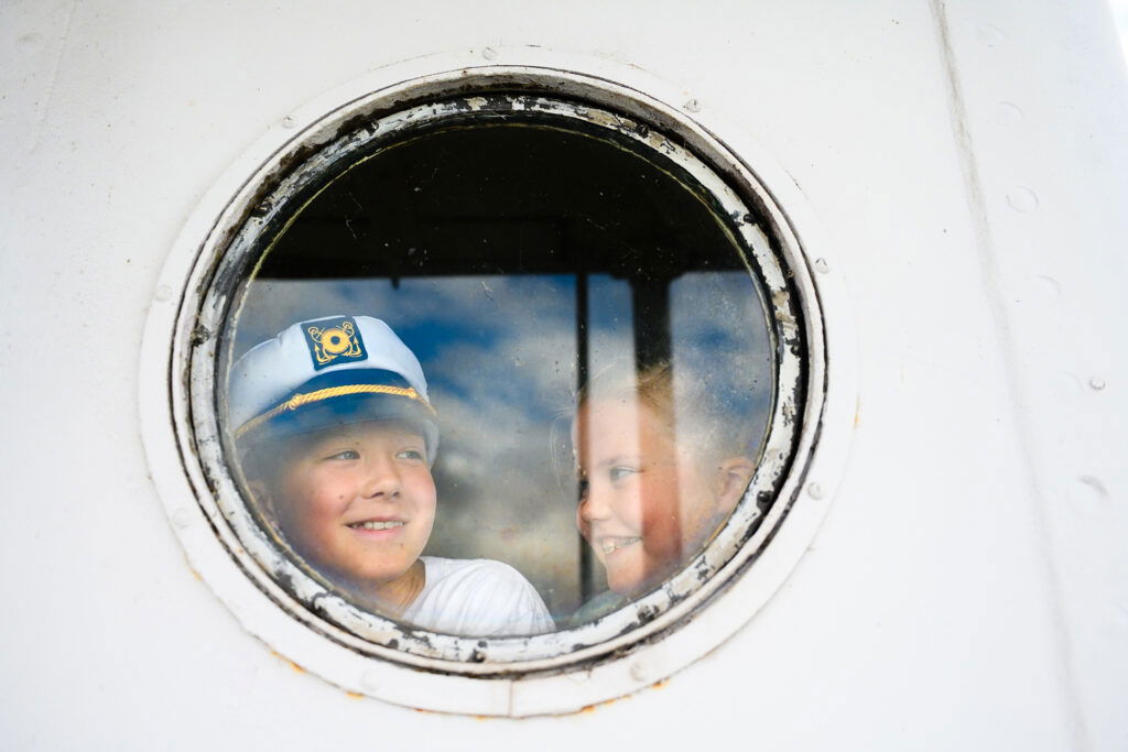 Two smiling children look out through a round ship window; one wears a captain's hat, both appear happy and playful, capturing the joy of a Wisconsin Family Vacation against the white frame and blue sky reflection.