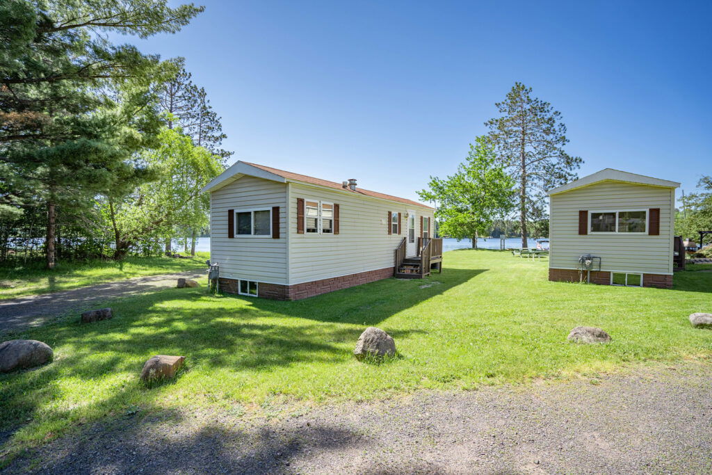 A light yellow mobile home with brown shutters and a nearby outbuilding sits on a grassy lawn, perfect for summer activities in Superior Wisconsin, surrounded by trees, with a lake and clear blue sky beyond. Rocks line the edge of a gravel driveway.