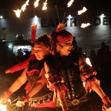 Two women dressed in elaborate costumes perform a fire dance at night, spinning flaming batons with bright flames illuminating their faces against a dark background.